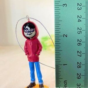 May include: A red and blue plastic figurine of Miguel from the movie Coco. The figurine is about 4 inches tall and is holding his hands in his pockets. The figurine is standing on a yellow base.