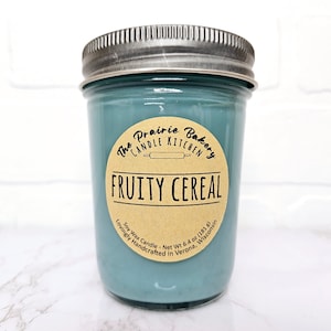 May include: A blue glass jar candle with a brown label that reads "The Prairie Bakery Candle Kitchen" and "Fruity Cereal". The label also includes the text "Soy Wax Candle - Net Wt 6.4 oz (181 g) Lovingly Handcrafted in Verona, Wisconsin".