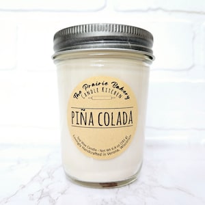 May include: A glass jar candle with a brown label that reads "The Prairie Bakery Candle Kitchen" and "PIÑA COLADA". The label also includes the text "Soy Wax Candle - Net Wt 6.4 oz (181 g) Lovingly Handcrafted in Verona, Wisconsin".