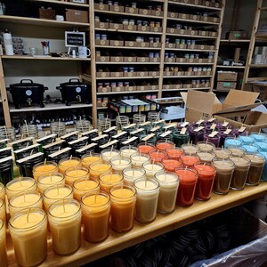May include: A table filled with rows of candles in various colors, including green, black, white, orange, red, brown, and blue. The candles are in glass jars with wicks and are arranged in a neat pattern.
