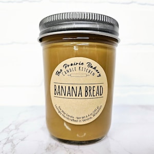 May include: A brown glass jar candle with a brown paper label that reads "The Prairie Bakery Candle Kitchen" and "Banana Bread". The label also includes the text "Soy Wax Candle - Net Wt 6.4 oz (181 g) Lovingly Handcrafted in Verona, Wisconsin".