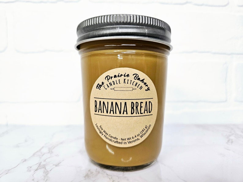 May include: A brown glass jar candle with a brown paper label that reads "The Prairie Bakery Candle Kitchen" and "Banana Bread". The label also includes the text "Soy Wax Candle - Net Wt 6.4 oz (181 g) Lovingly Handcrafted in Verona, Wisconsin".