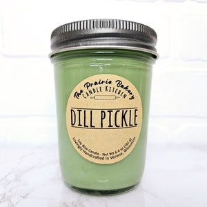 May include: A green soy wax candle in a glass jar with a brown label that reads "The Prairie Bakery Candle Kitchen Dill Pickle Lovingly Handcrafted in Verona, Wisconsin Soy Wax Candle - Net Wt 6.4 oz (181 g)"
