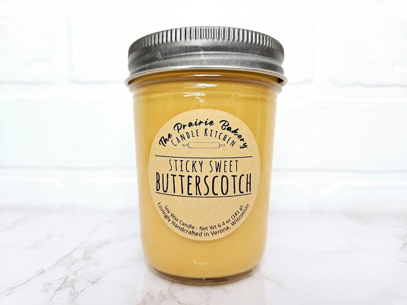 May include: A jar candle with a silver lid filled with a light amber wax. The label reads "Sticky Sweet Butterscotch" from The Prairie Bakery Candle Kitchen. The candle is 6.4 oz (181 g) and made with soy wax.