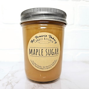 May include: A brown glass jar candle with a brown paper label that reads "The Prairie Bakery Candle Kitchen Maple Sugar Soy Wax Candle - Net Wt 6.4 oz (181 g) Lovingly Handcrafted in Verona, Wisconsin"
