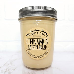 May include: A brown glass jar candle with a brown paper label that reads "The Prairie Bakery Candle Kitchen" and "Cinnamon Raisin Bread". The label also includes the text "Soy Wax Candle - Net Wt 6.4 oz (181 g) Lovingly Handcrafted in Verona, Wisconsin".