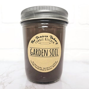 May include: A soy wax candle in a clear glass jar with a silver lid. The candle is dark brown and has a label that reads "Garden Soil" from The Prairie Bakery Candle Kitchen. Net weight is 6.4 oz (181 g).