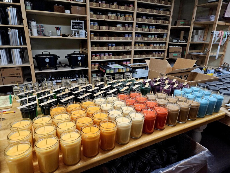 May include: A variety of jar candles in various colors, including orange, yellow, red, brown, green, purple, and blue, are arranged on a wooden table. The candles are in clear glass jars with wooden wicks. Shelves filled with supplies are in the background.