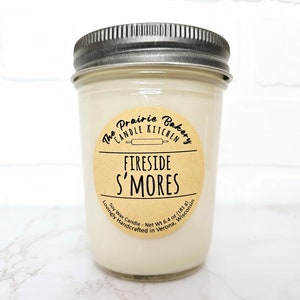May include: A glass jar candle with a brown label that reads "The Prairie Bakery Candle Kitchen" and "Fireside S'mores". The label also includes the text "Soy Wax Candle - Net Wt 6.4 oz (181 g) Lovingly Handcrafted in Verona, Wisconsin".