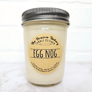 May include: A glass jar candle with a brown label that reads "The Prairie Bakery Candle Kitchen Egg Nog" and "Soy Wax Candle - Net Wt 6.4 oz (181 g) Lovingly Handcrafted in Verona, Wisconsin".