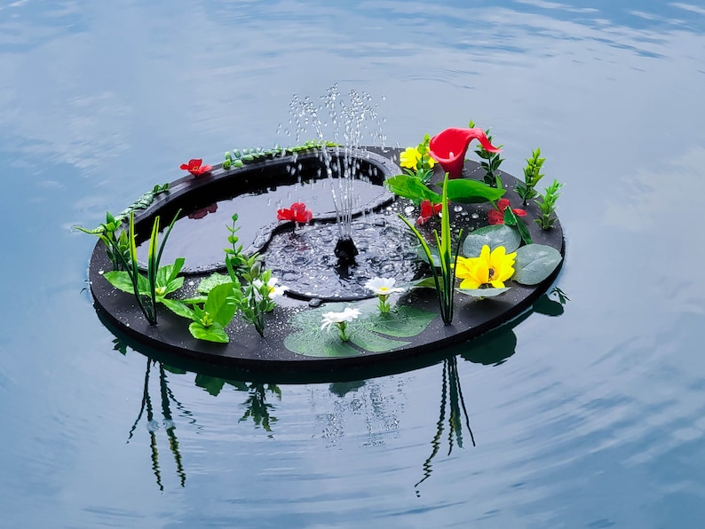 May include: A black floating fountain with a central water jet, surrounded by artificial flowers and greenery. The fountain features red, yellow, and white flowers, and is set against a backdrop of blue water.