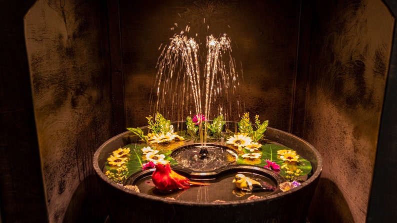 May include: A decorative fountain with water jets, surrounded by flowers and artificial birds. The fountain is set in a dark, wooden barrel-like structure, creating a serene and natural ambiance. The water is illuminated, enhancing the visual appeal.