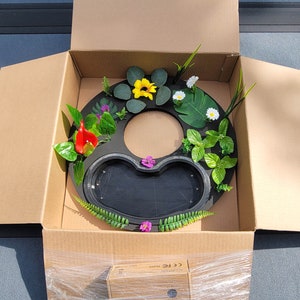 May include: A black, circular fountain base with a heart-shaped basin, decorated with artificial flowers and greenery, including yellow sunflowers, red calla lilies, and white daisies. The fountain is packaged in a cardboard box.
