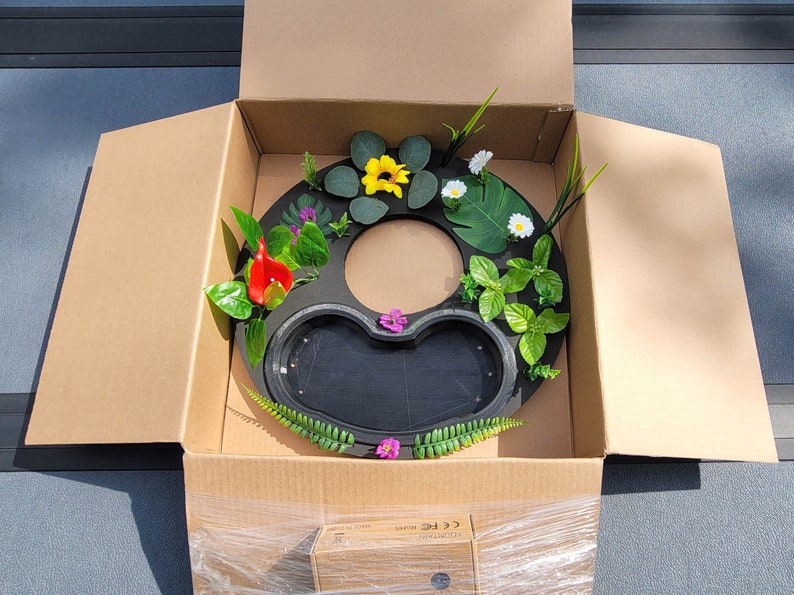 May include: A black, circular fountain base with a heart-shaped basin, decorated with artificial flowers and greenery, including yellow sunflowers, red calla lilies, and white daisies. The fountain is packaged in a cardboard box.