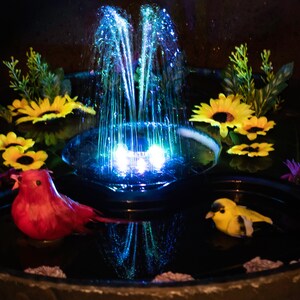 May include: A decorative water fountain with a central water jet illuminated by blue lights. The fountain is surrounded by yellow sunflowers, green foliage, and two bird figurines. The fountain is in a dark setting.