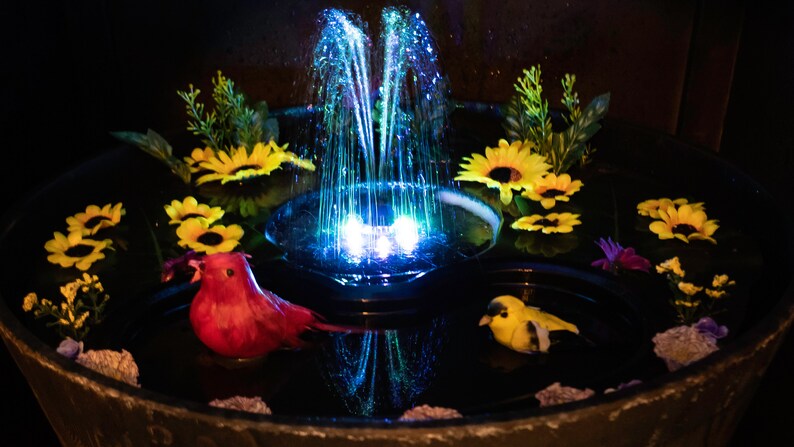 May include: A decorative water fountain with a central water jet illuminated by blue lights. The fountain is surrounded by yellow sunflowers, green foliage, and two bird figurines. The fountain is in a dark setting.