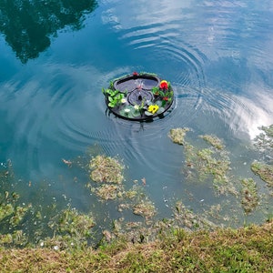 May include: A floating pond fountain with a black base, adorned with colorful artificial flowers and greenery. The fountain sprays water into the air, creating ripples on the water's surface. The pond water is a deep teal color.