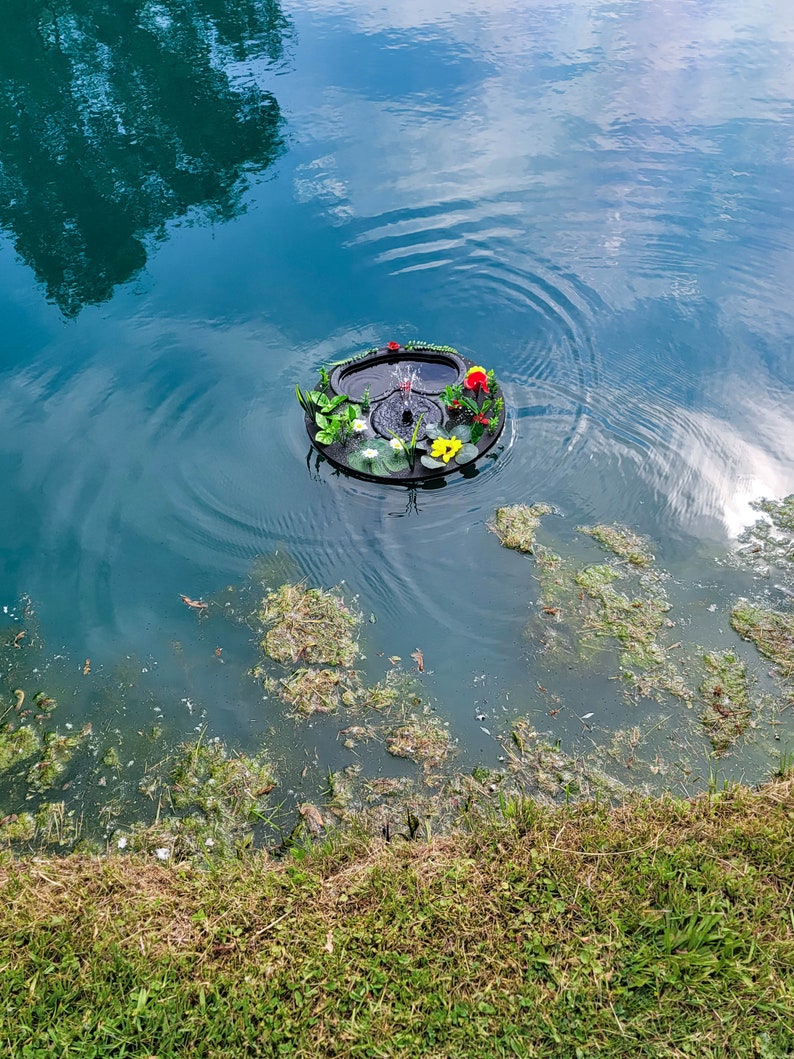 May include: A floating pond fountain with a black base, adorned with colorful artificial flowers and greenery. The fountain sprays water into the air, creating ripples on the water's surface. The pond water is a deep teal color.