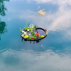 May include: A floating miniature pond with a black base, surrounded by colorful artificial flowers and greenery. The pond features a small fountain and is set against a backdrop of blue water with reflections of the sky.