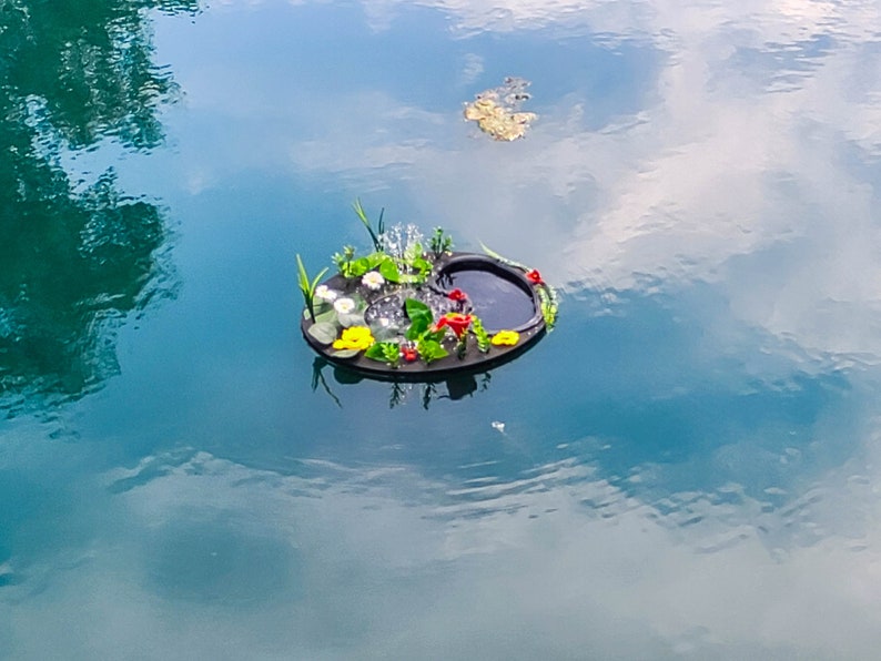 May include: A floating miniature pond with a black base, surrounded by colorful artificial flowers and greenery. The pond features a small fountain and is set against a backdrop of blue water with reflections of the sky.