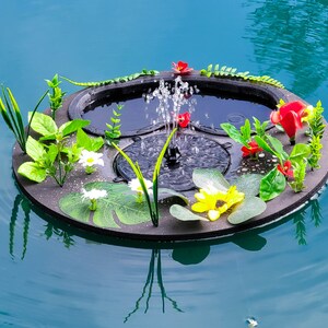 May include: A floating fountain with a black base, surrounded by artificial flowers and greenery. The fountain sprays water upwards, creating a small water feature. The flowers include red, yellow, and white blooms, with green leaves.