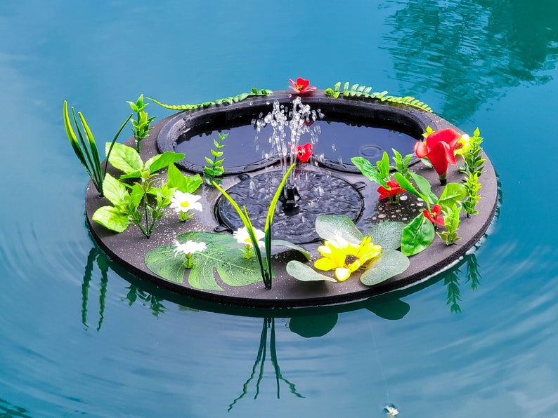 May include: A floating fountain with a black base, surrounded by artificial flowers and greenery. The fountain sprays water upwards, creating a small water feature. The flowers include red, yellow, and white blooms, with green leaves.