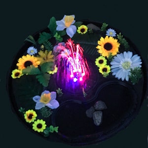 May include: A decorative, round fountain with a black base, surrounded by colorful artificial flowers, including sunflowers and daisies. The fountain has a central water feature with red and purple lights, and small rocks.
