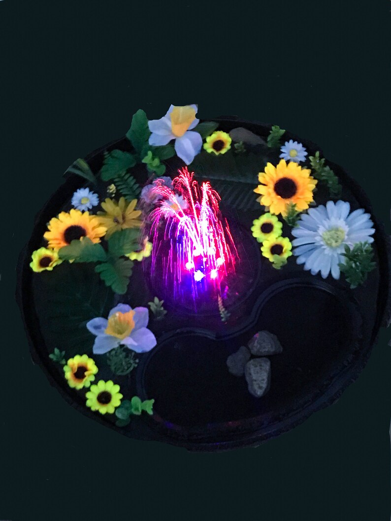 May include: A decorative, round fountain with a black base, surrounded by colorful artificial flowers, including sunflowers and daisies. The fountain has a central water feature with red and purple lights, and small rocks.