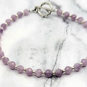 May include: A delicate bracelet featuring faceted, lavender-colored beads strung together with small silver-toned beads. The bracelet is secured with a silver toggle clasp. The beads have a subtle sparkle, and the overall design is elegant and refined.