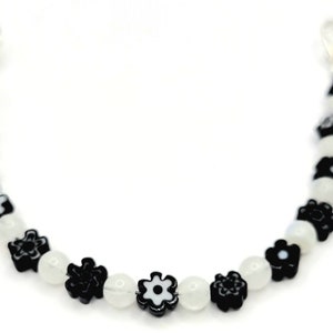 Op de afbeelding: Een armband met afwisselend witte ronde kralen en zwarte bloemvormige kralen. De armband heeft zilverkleurige sluitingen aan elk uiteinde. De bloemen hebben witte centra en zwarte bloemblaadjes. Op een witte achtergrond.