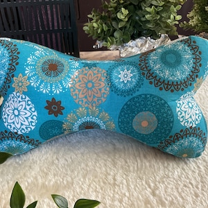 Op de afbeelding: Een turquoise blauw botvormig kussen met een mandala-patroon in tinten bruin, wit en goud.