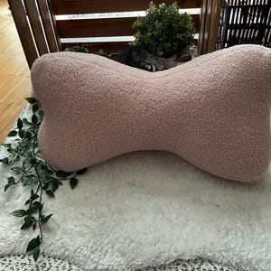 Op de afbeelding: Een lichtroze, botvormig kussen gemaakt van een zacht, getextureerd materiaal. Het kussen ligt op een wit, pluizig tapijt, met groene, bladrijke wijnranken op de voorgrond. Het kussen is ontworpen voor nekondersteuning.