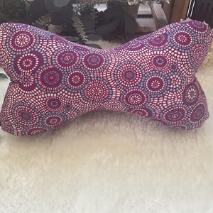 Op de afbeelding: Een decoratief botvormig kussen met een herhalend patroon van paarse, witte en lichtblauwe cirkels. Het kussen is gemaakt van stof en ligt op een witte, zachte ondergrond. Het ontwerp van het kussen is symmetrisch, met afgeronde uiteinden en een gebogen midden.