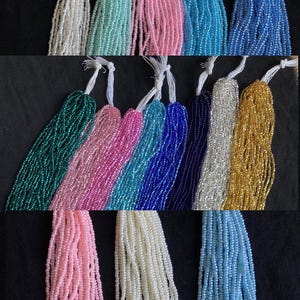 Puede incluir: Surtido de cuentas de semillas en varios colores, incluyendo blanco, azul claro, rosa, verde azulado, dorado y plateado. Las cuentas están dispuestas en filas, con cada fila que contiene un color diferente. Las cuentas son todas del mismo tamaño y forma.