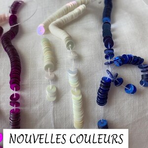 May include: Four strands of sequins in different colors: pink, purple, white, and blue. The sequins are strung on a clear thread and are arranged in a line. The text "NOUVELLES COULEURS" is written below the sequins.