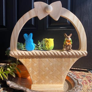 May include: A wooden Easter basket with a bow on top. The basket is decorated with a rope trim and a woven pattern. It is filled with a blue bunny, a yellow tulip, and a brown bunny holding a carrot.