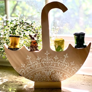 May include: A wooden umbrella-shaped stand with a decorative white design. It features three small ceramic figurines: a rabbit, a pair of yellow rain boots, and a pair of green rain boots.