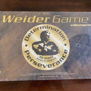 Peut inclure: La boîte de jeu de société Weider Game présente un cercle doré avec les mots "Détermination, Persévérance" et une image en noir et blanc de Joe Weider, "Entraîneur des champions". La boîte est brune avec un fond doré et le texte "C'est plus qu'un jeu !"