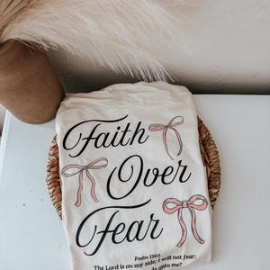May include: Cream-colored t-shirt with the words "Faith Over Fear" in elegant script, accented with pink bow illustrations. The shirt also features a Bible verse. The shirt is displayed on a woven basket.
