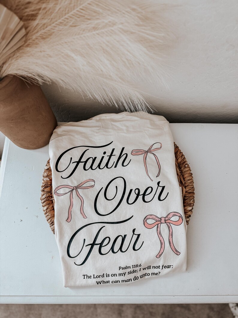 May include: Cream-colored t-shirt with the words "Faith Over Fear" in elegant script, accented with pink bow illustrations. The shirt also features a Bible verse. The shirt is displayed on a woven basket.