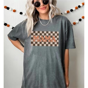 May include: Gray t-shirt with the word "MAMA" in a peach-colored, retro font, set against a brown and tan checkered background. The shirt has short sleeves and a relaxed fit. A decorative garland hangs in the background.
