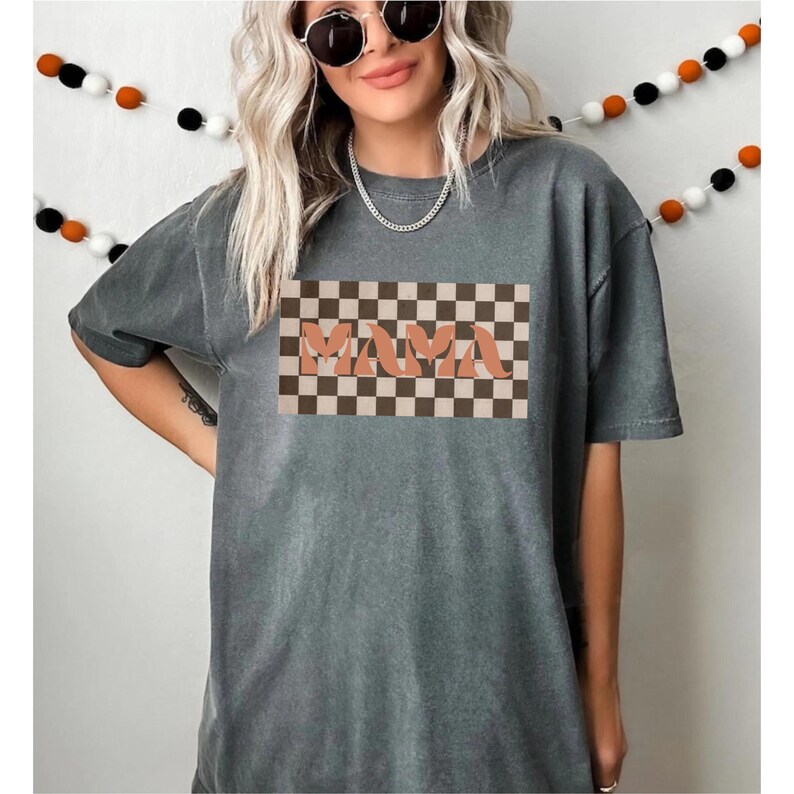 May include: Gray t-shirt with the word "MAMA" in a peach-colored, retro font, set against a brown and tan checkered background. The shirt has short sleeves and a relaxed fit. A decorative garland hangs in the background.