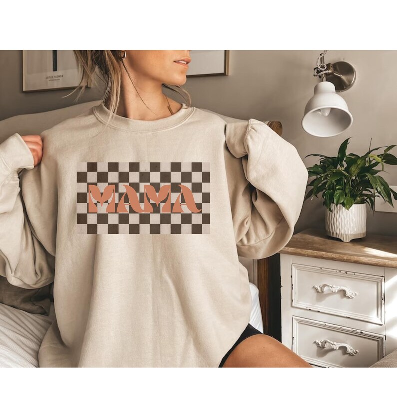 May include: Beige sweatshirt with a retro-style checkered design and the word "MAMA" in a peach-colored font. The sweatshirt has long sleeves and a crew neck. The design is centered on the front of the shirt.