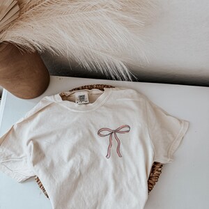 May include: Cream-colored t-shirt with a pink bow graphic on the front. The shirt is folded on a woven basket. The shirt has a tag at the neckline. A brown vase and dried plants are in the background.