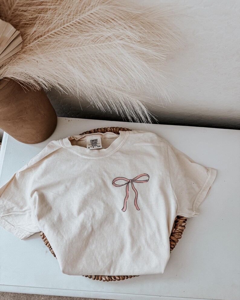 May include: Cream-colored t-shirt with a pink bow graphic on the front. The shirt is folded on a woven basket. The shirt has a tag at the neckline. A brown vase and dried plants are in the background.