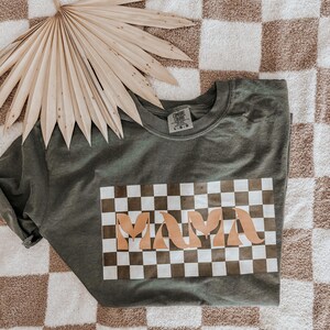 May include: Olive green crew neck t-shirt with a checkered design and the word "MAMA" in a light brown color. The shirt is folded on a checkered brown and white surface, with a dried palm leaf in the background.