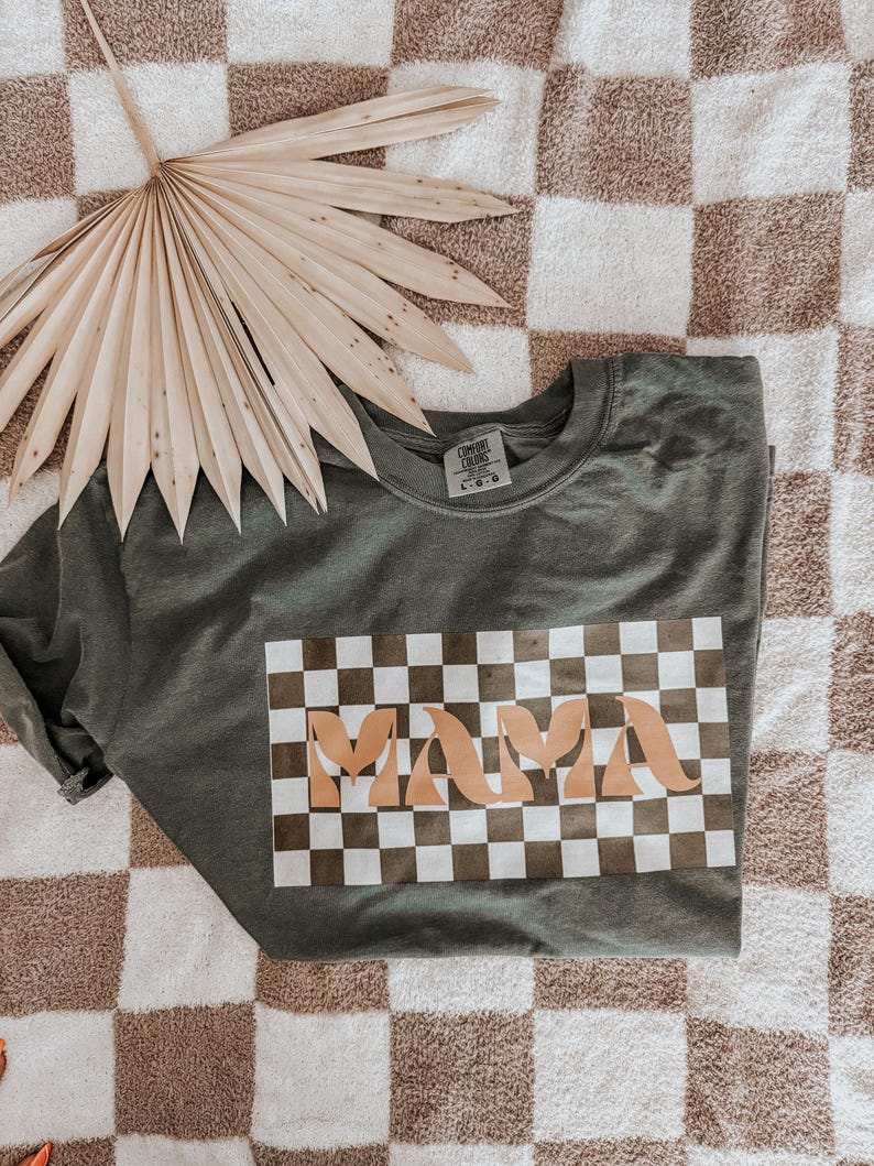 May include: Olive green crew neck t-shirt with a checkered design and the word "MAMA" in a light brown color. The shirt is folded on a checkered brown and white surface, with a dried palm leaf in the background.
