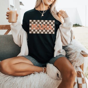May include: Black t-shirt with a retro checkered design and the word "MAMA" in a peach-colored font. The shirt is part of a casual clothing collection. The model is wearing a cream-colored cardigan and denim shorts.