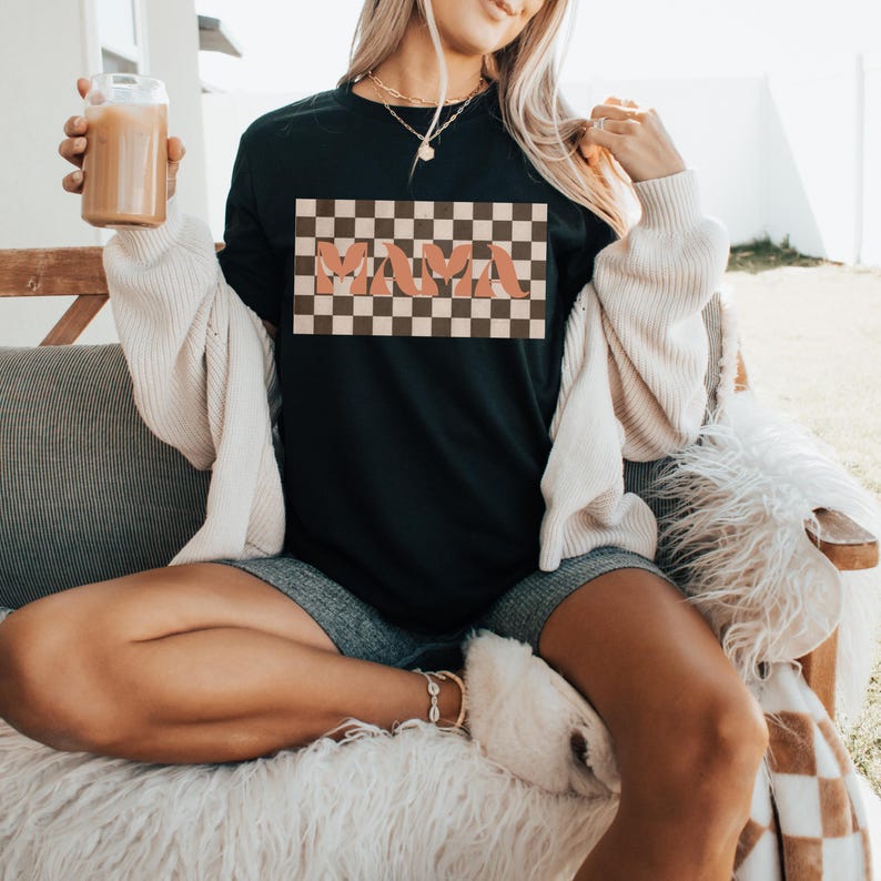 May include: Black t-shirt with a retro checkered design and the word "MAMA" in a peach-colored font. The shirt is part of a casual clothing collection. The model is wearing a cream-colored cardigan and denim shorts.