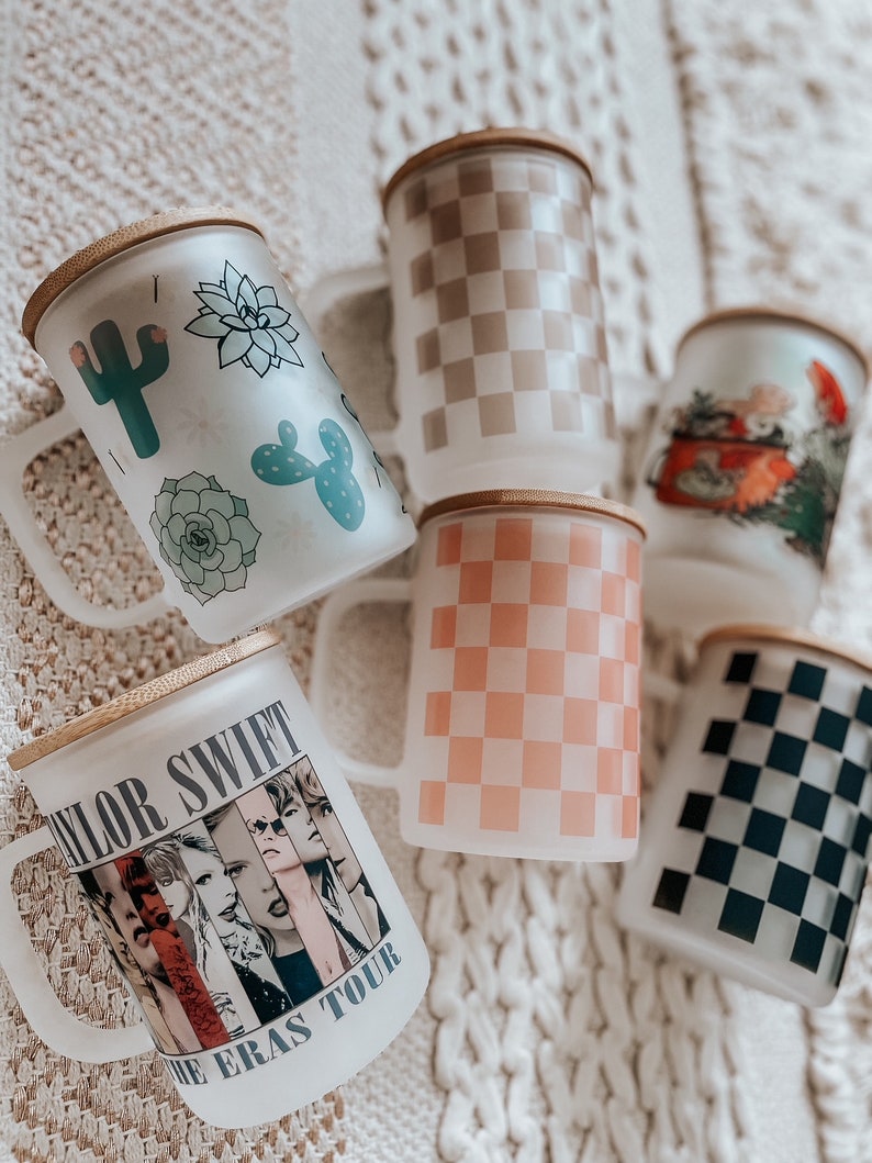 May include: Six different ceramic mugs with wooden lids. The mugs feature various designs, including a checkered pattern, a cactus and succulent design, a car design, and a Taylor Swift "The Eras Tour" design.